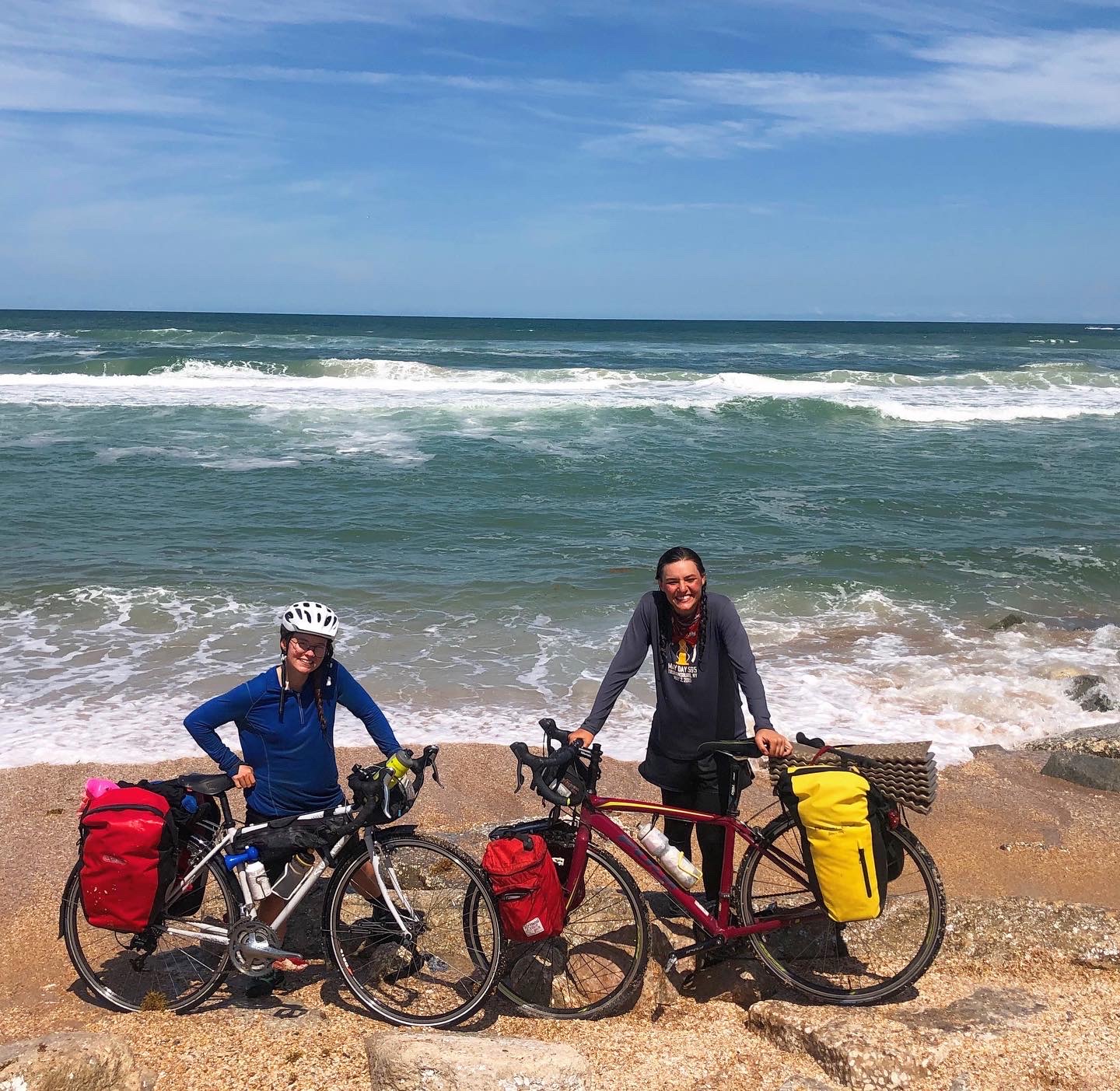 Two smiling women pose with bicycles on a beach near the waves