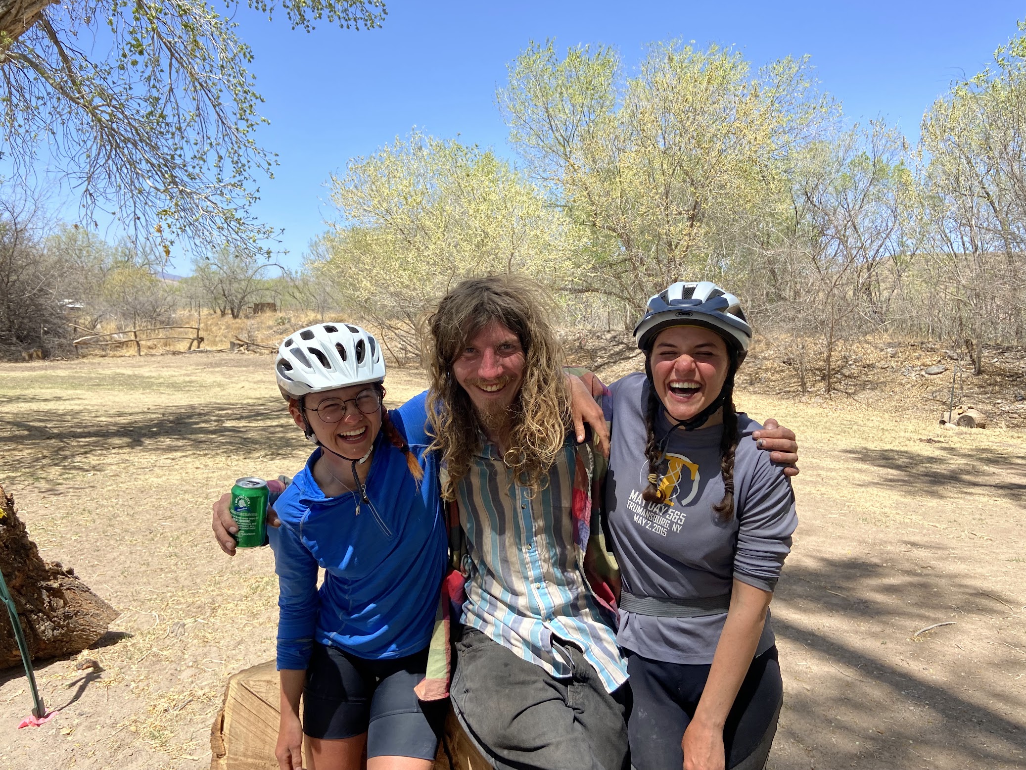 Two laughing women wearing bicycle helmets hug the shoulders of a smiling man with long blonde hair